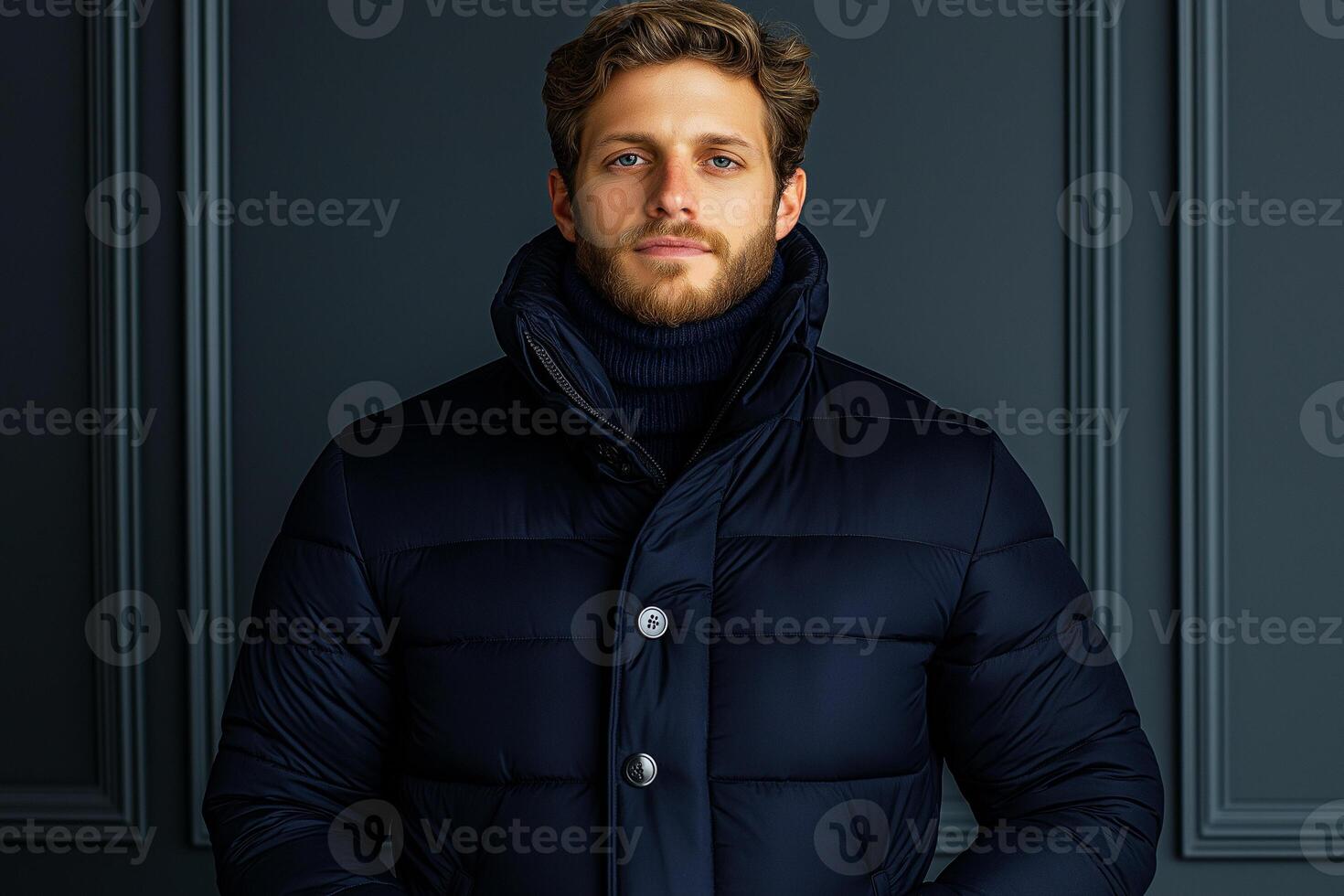 man in stylish navy puffer jacket stands confidently against dark wall. His expression is calm and composed, showcasing modern fashion and elegance photo