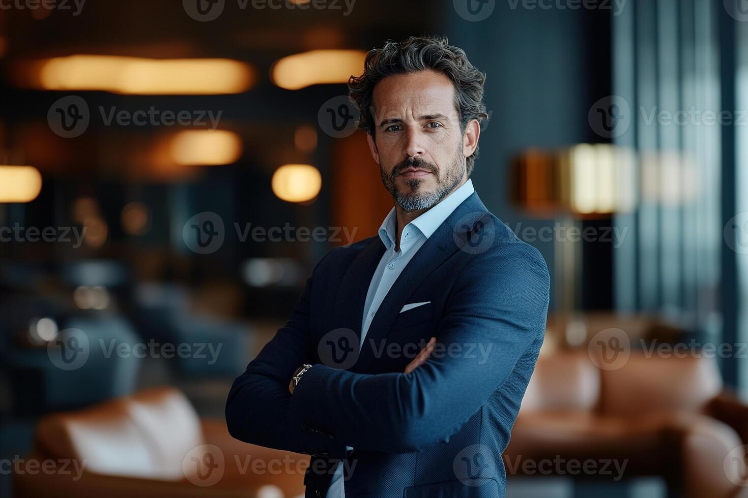 The confident man stands with arms crossed in a modern lounge, wearing a tailored navy-blue suit. Soft golden lights reflect off the leather furniture, creating a stylish atmosphere photo