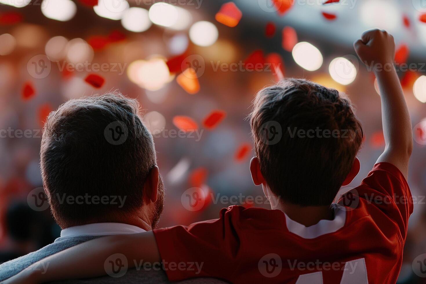 A father and son celebrate together at a crowded football stadium. They wear matching jerseys and cheer passionately as the crowd roars around them. The soft lighting creates a vibrant atmosphere photo