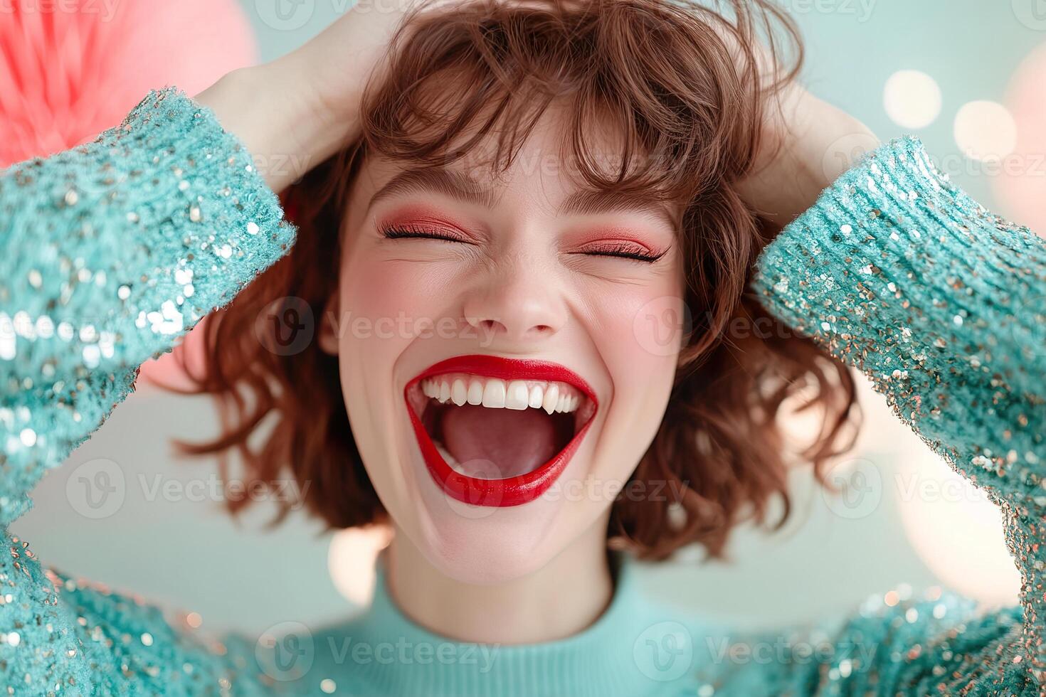 A fashionable woman with bold red lipstick, smiling broadly and laughing. Her hands are playfully placed on her head, eyes closed, against a blurred pastel colored backdrop photo