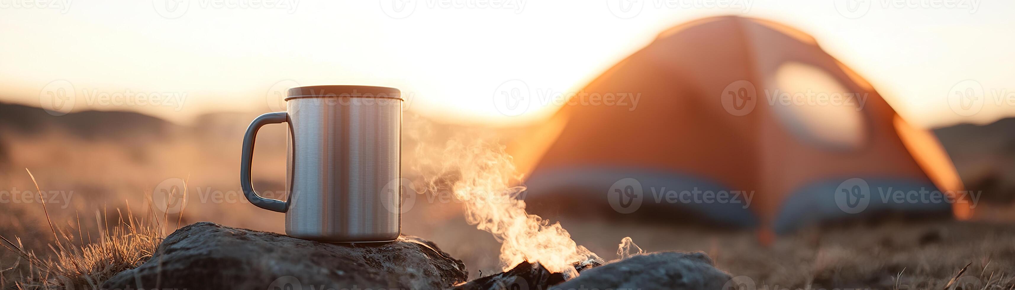 Tucked away in a remote field, a small, inexpensive camping tent rises against the soft morning light. Nearby, a steaming metal mug sits on a rock, alongside a gentle, smoldering fire photo