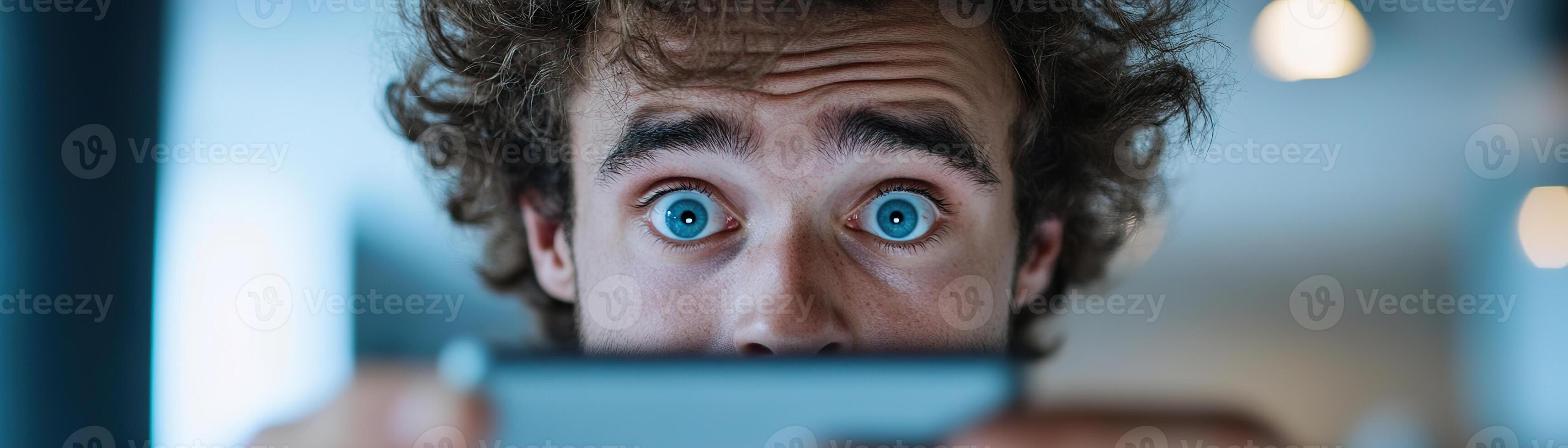A young man with curly hair holds his smartphone up, gazing at the screen with wide eyes and an open mouth, clearly astonished by what he sees in a soft-focus techy background photo