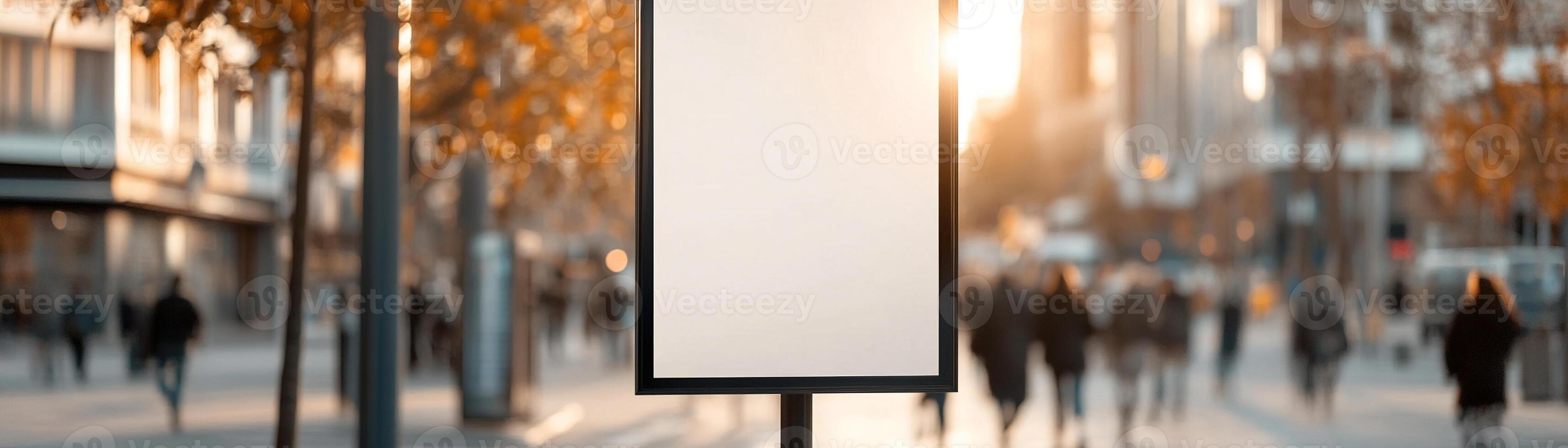 A large blank banner hangs from a black lamppost in a bustling downtown street. Sunlight filters through trees, casting gentle shadows as pedestrians stroll by, creating a vibrant urban atmosphere photo