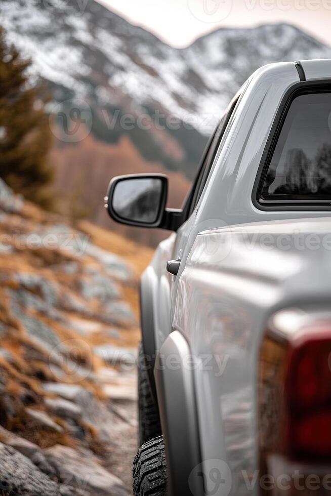 A silver truck is parked on a rugged mountain trail surrounded by rocky terrain and autumn foliage, with snow-capped peaks visible in the distance under clear skies photo
