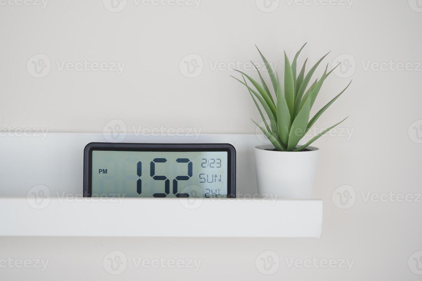 Digital clock on shelf next to small potted plant in afternoon light photo