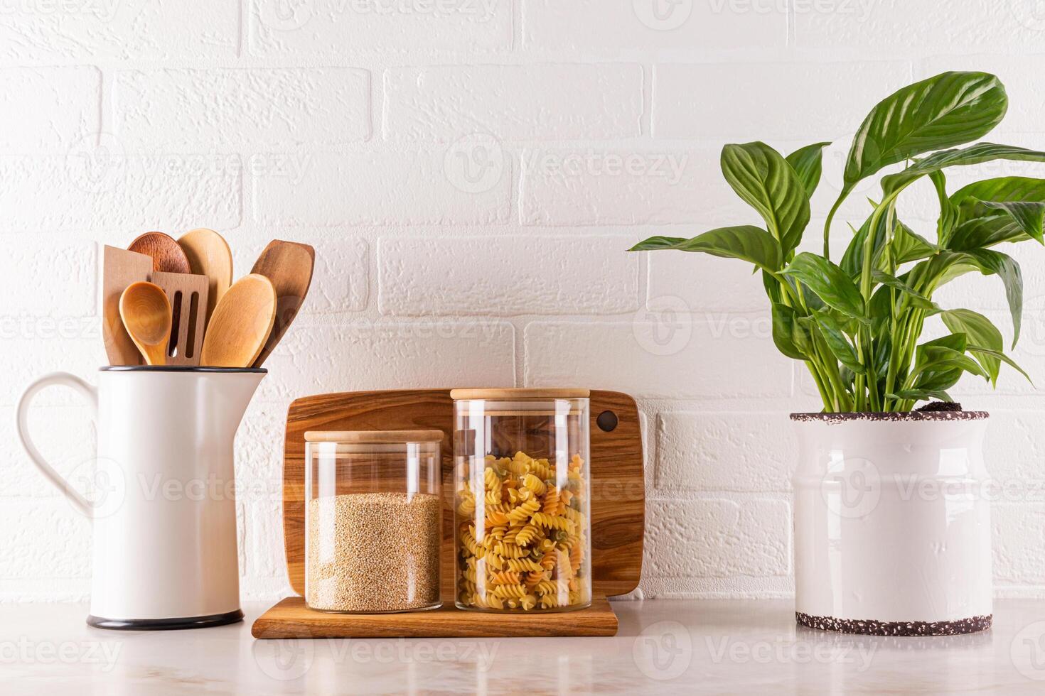 Stylish glass jars for storing bulk products, jug with wooden spoons, beautiful indoor flower in a modern pot on a marble countertop. Front view. photo