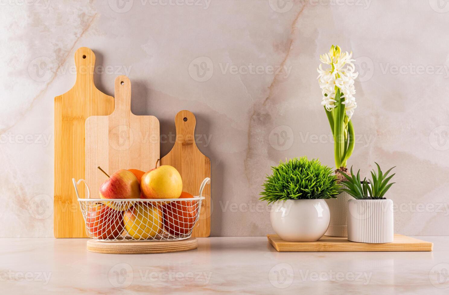 Chic kitchen still life with a set of cutting wooden boards, indoor flowering plants and a wicker fruit bowl on a marble beige countertop. Front view. photo