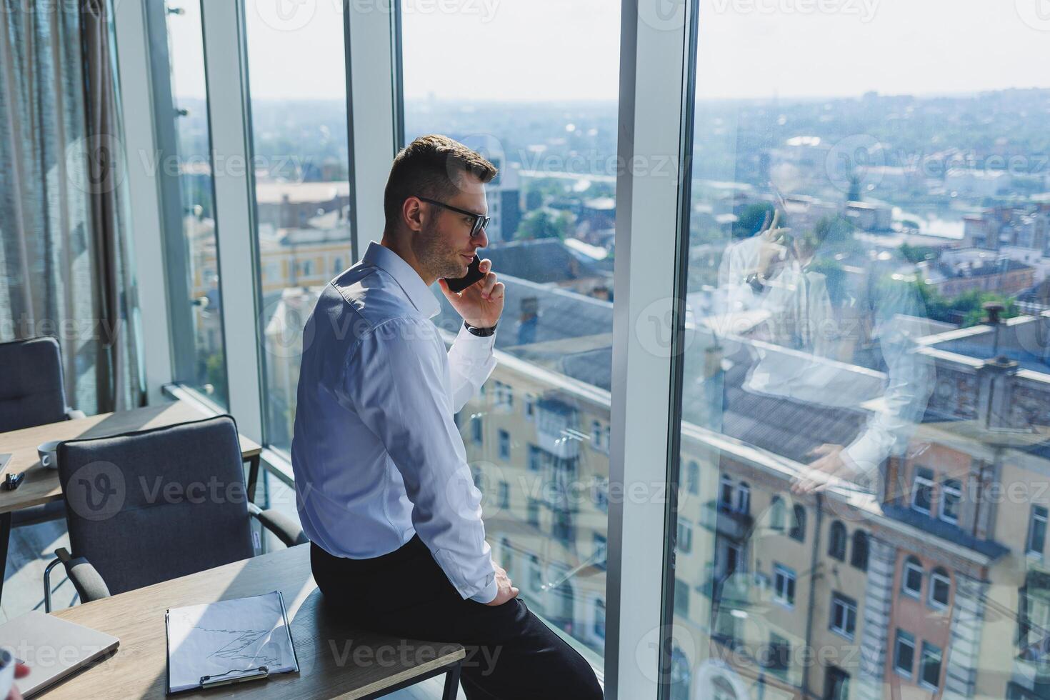 A young businessman with a phone in his hands in glasses and a white shirt, he is in a modern office with large windows. A man in classic trousers and a white shirt. Young attractive businessman photo