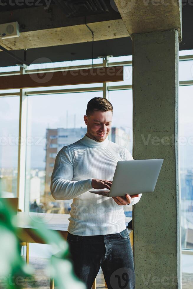 A young man in a modern workspace stands near a large window. A man in casual clothes with a laptop in his hands. Remote work. Nice young manager photo