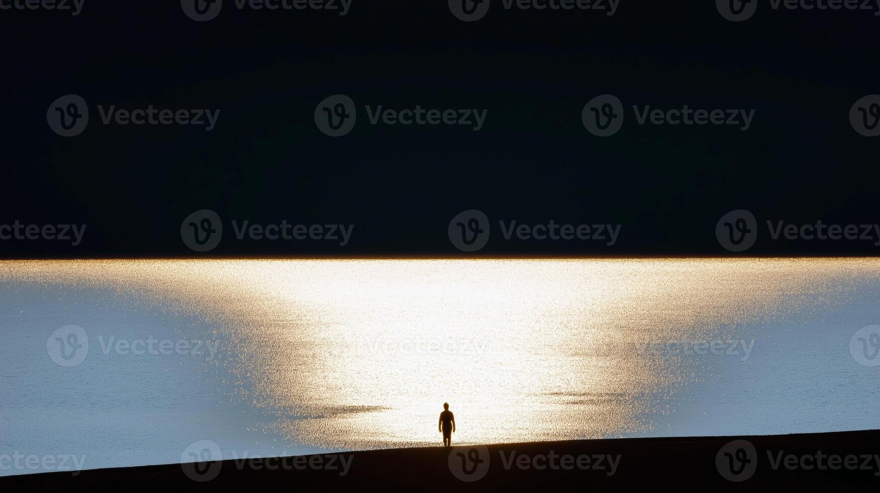 Silhouette of a person standing near a calm lake with golden sunset reflection creating a serene and dramatic contrast photo