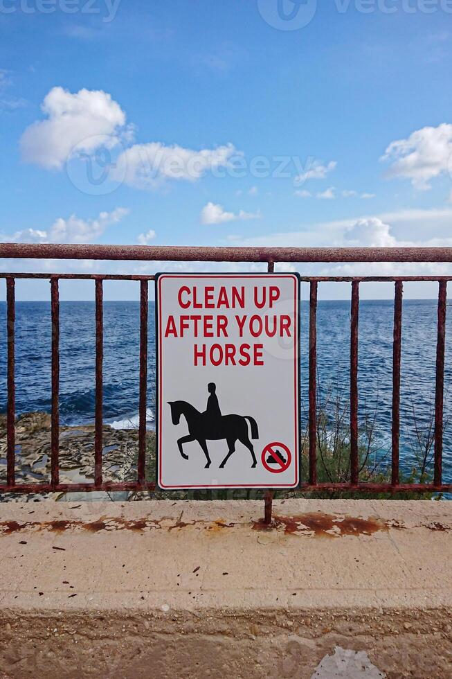 Sign instructing horse riders to clean up after their horses is mounted on rusted metal railing along scenic coastal pathway photo