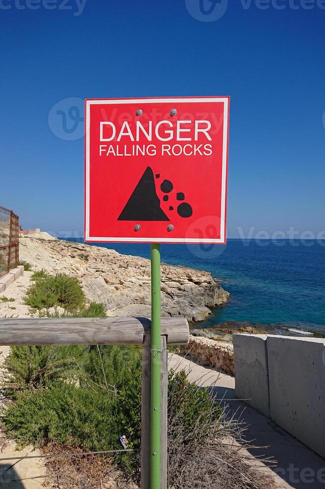 Bright red danger sign warns of falling rocks along scenic coast, danger emphasizing sudden hazards near cliffs, danger prompting cautious steps in public walkway by sea photo