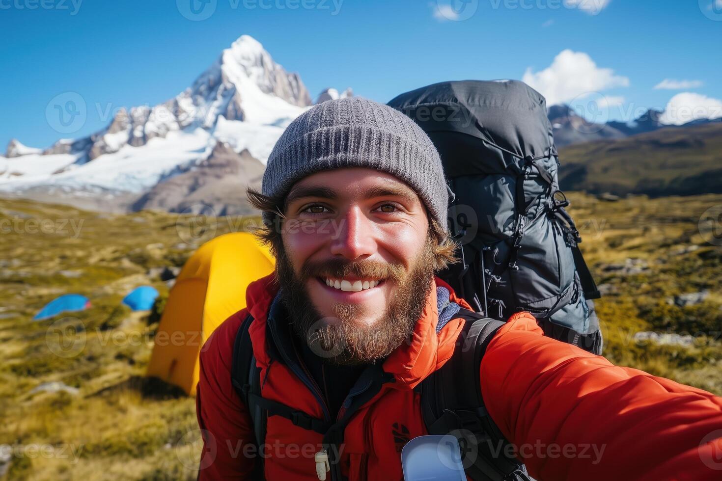 Smiling backpacker taking selfie with snowy mountain backdrop and camping tents in scenic outdoor adventure setting photo