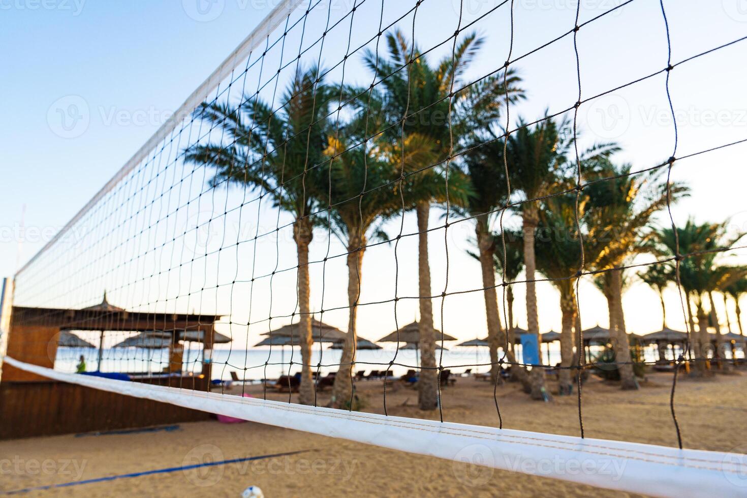 Volleyball net stands on sandy beach with palm trees and distant umbrellas during sunset photo