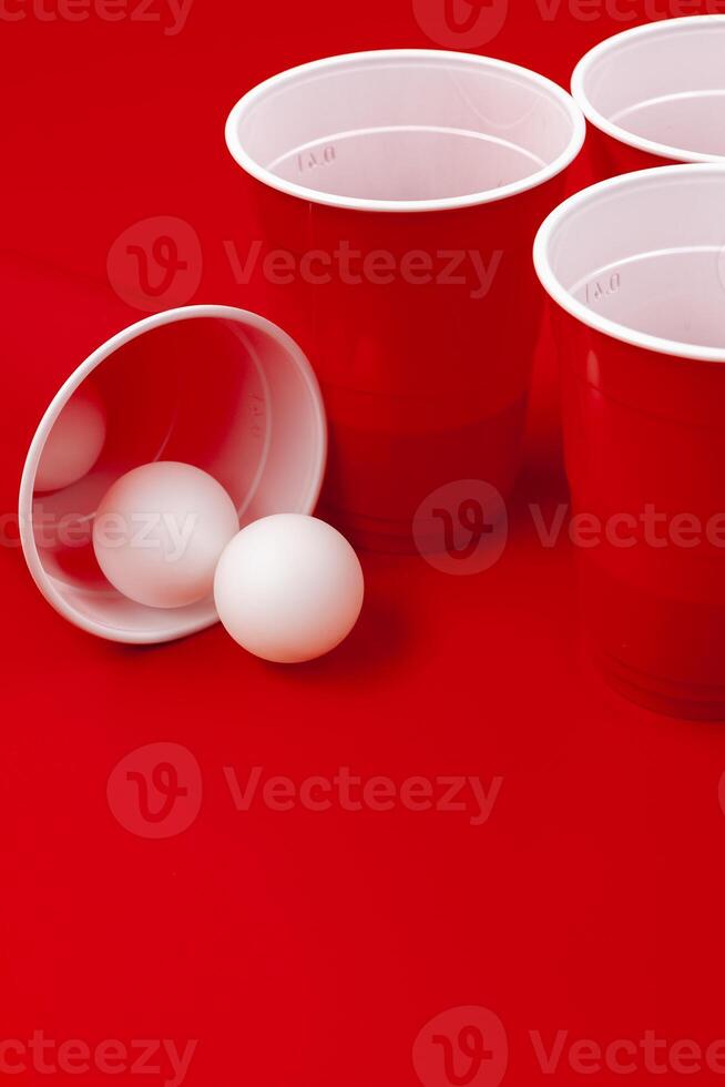 Cups and plastic ball on red background. Beer pong game photo