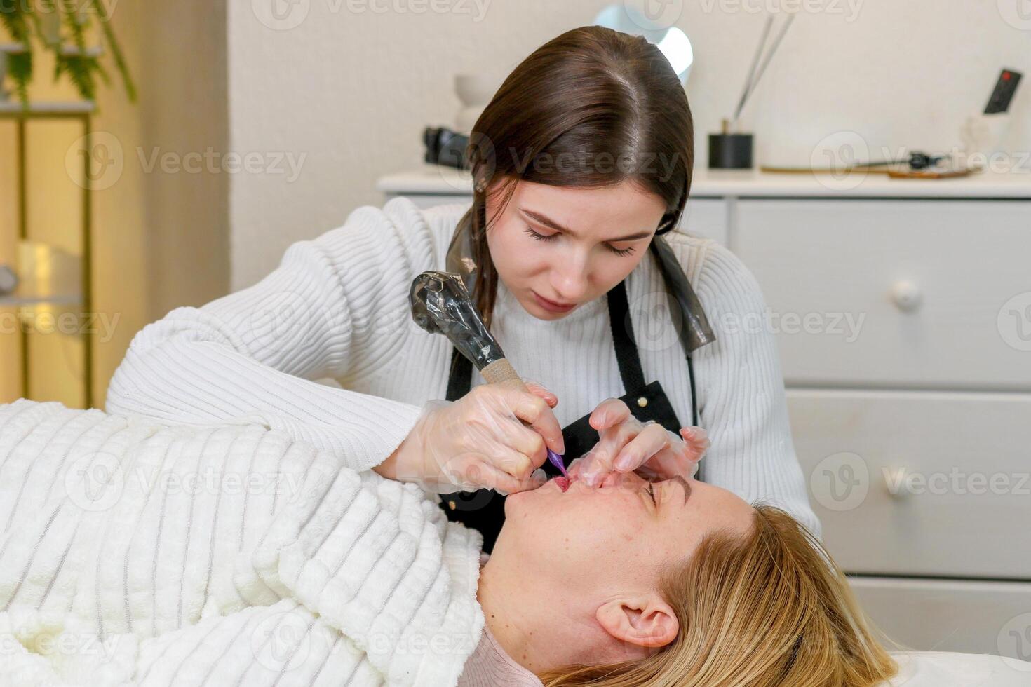 girl permanent makeup artist doing permanent lip makeup to middle aged woman photo