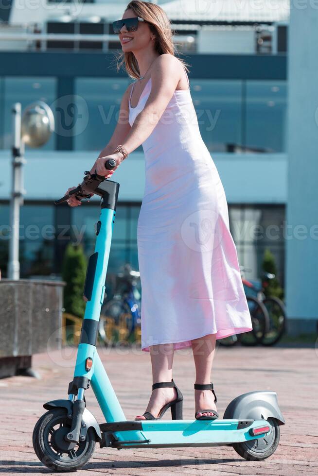 Elegant woman in a pink dress and sunglasses riding an electric scooter through the sunny city streets, embracing the freedom and style of a perfect urban adventure. photo