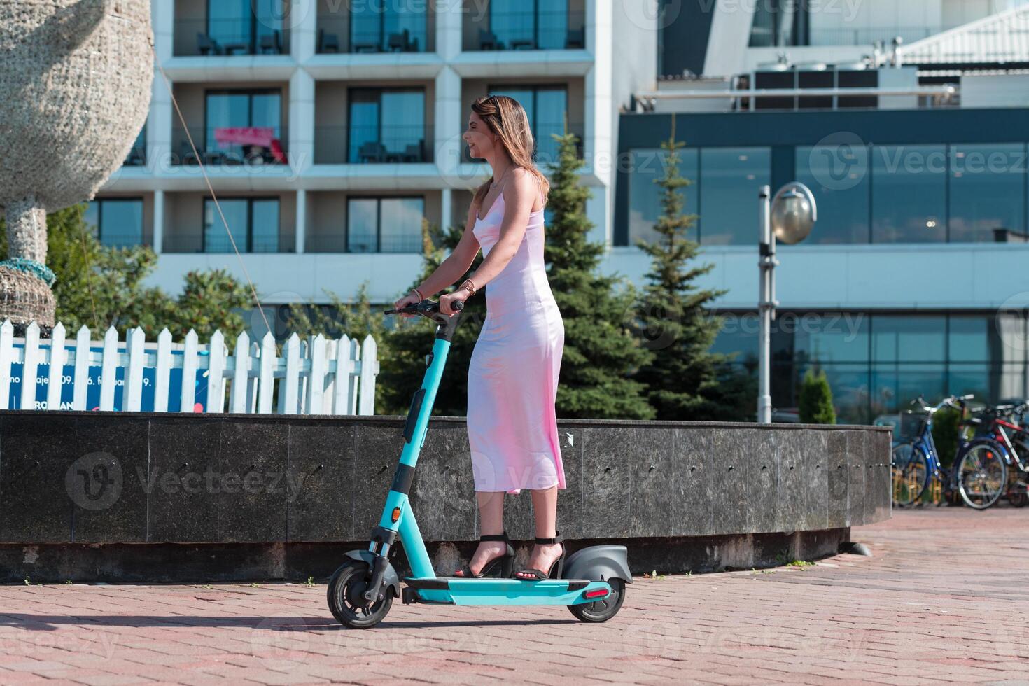 Elegant woman in a pink dress and sunglasses riding an electric scooter through the sunny city streets, embracing the freedom and style of a perfect urban adventure. photo