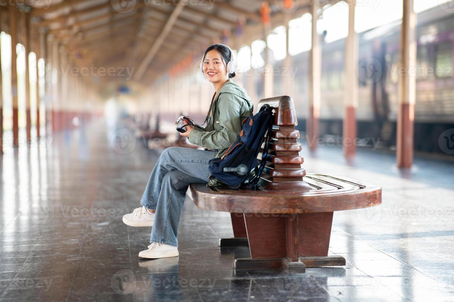 Smiling asian woman is holding her camera while sitting cross legged on the bench in train station. photo
