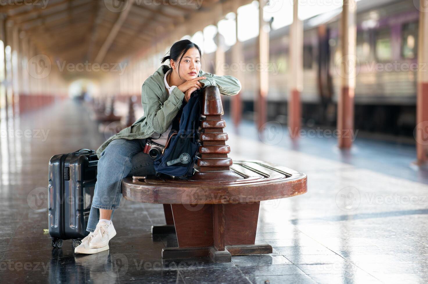 An asian woman is looking bored while sitting on wooden bench with her belonging in train station. photo