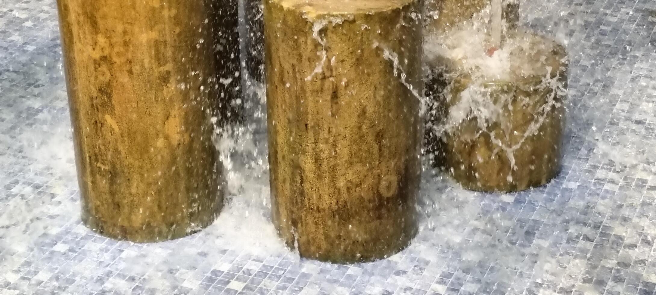 Water cascades over cylindrical wooden stumps in a fountain, creating dynamic splashes. The stumps stand on a mosaic-tiled surface, enhancing the rustic-meets-modern aesthetic of the water feature. photo