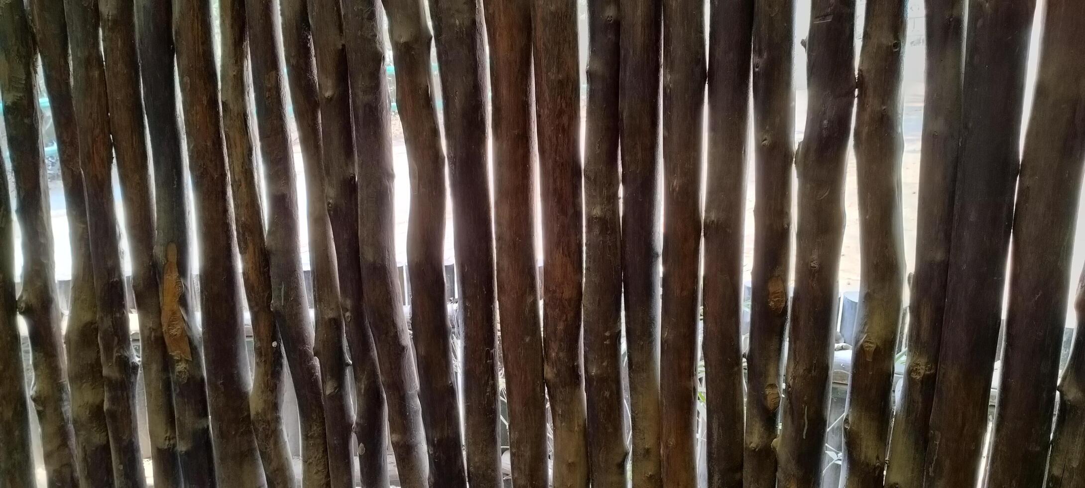 A rustic wooden fence made of vertical sticks, irregularly placed, allows glimpses of light and the world beyond. The natural textures and earthy tones create a raw, organic, and warm aesthetic. photo