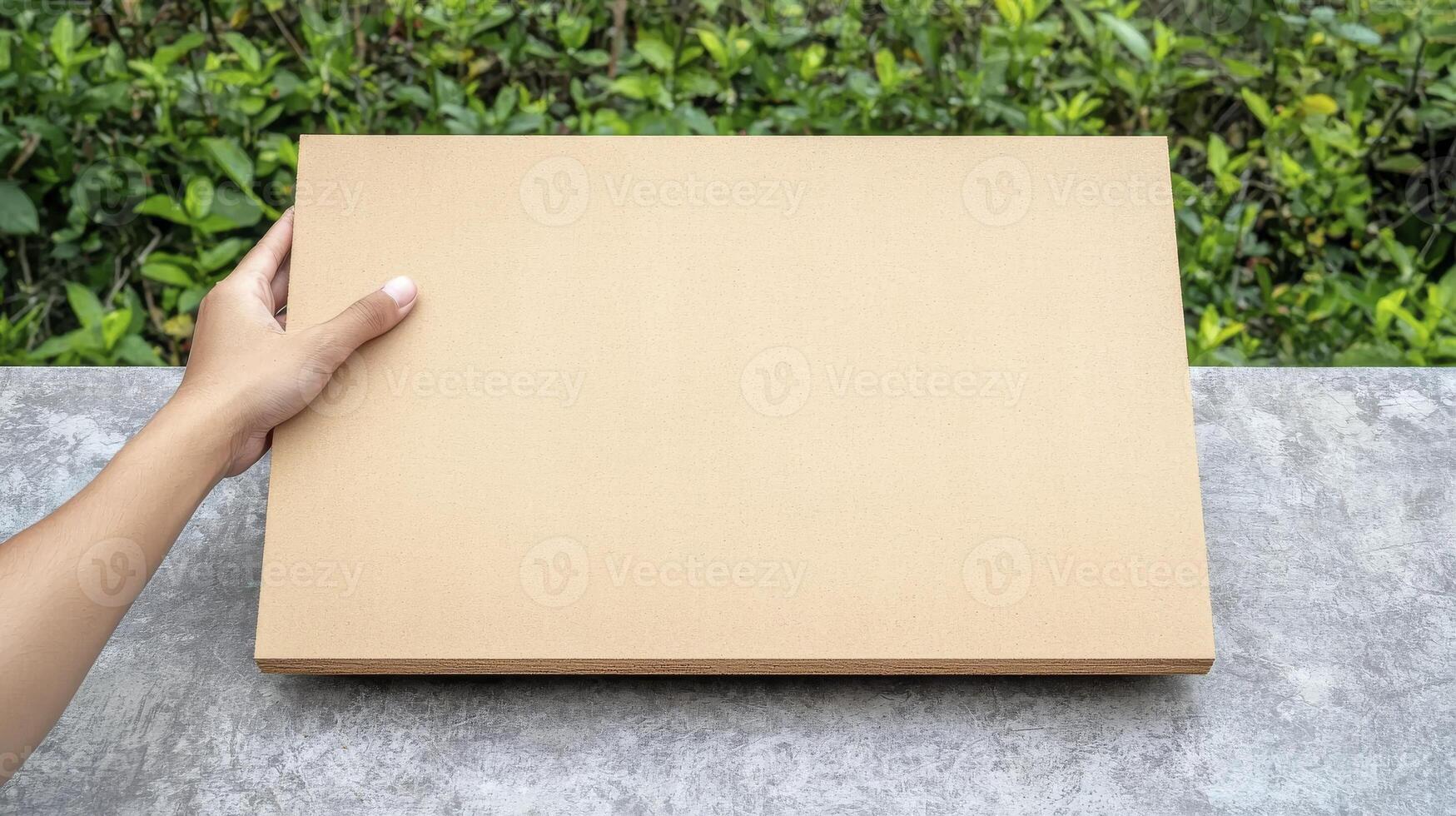 Hand Holding Empty Craft Paper Board Over Concrete Surface Surrounded by Green Foliage in Natural Outdoor Setting photo