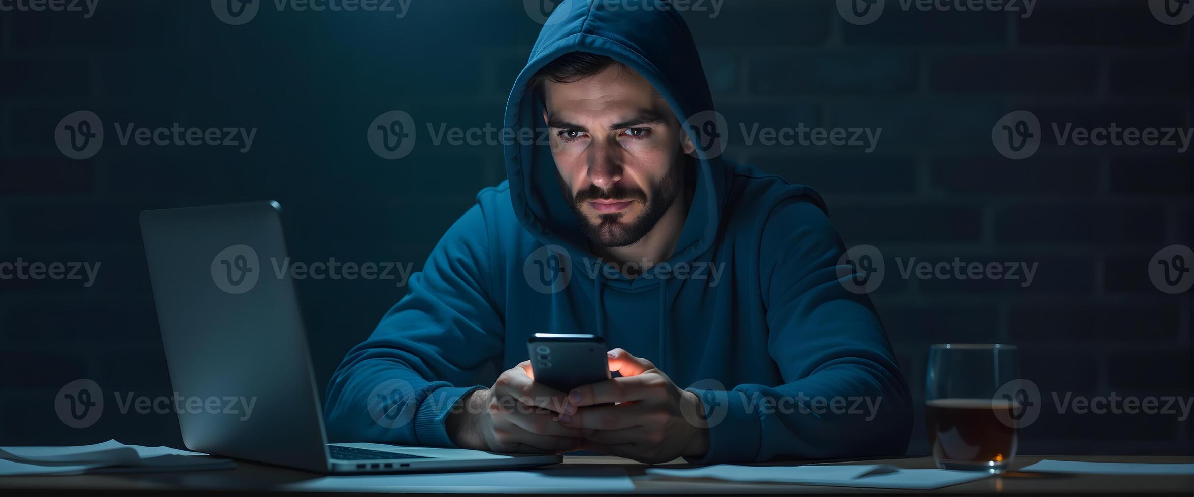 Man In Hoodie Using Smartphone With Laptop on Desk photo