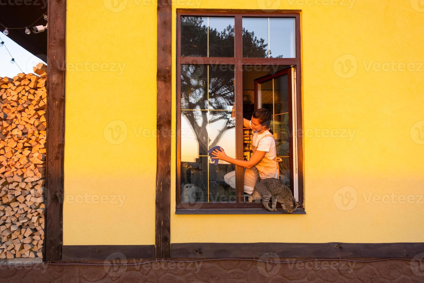 Woman in apron manually washes the window of the house with a rag with a spray cleaner and a mop outside.at height, restoring order and cleanliness in the spring, cleaning service photo