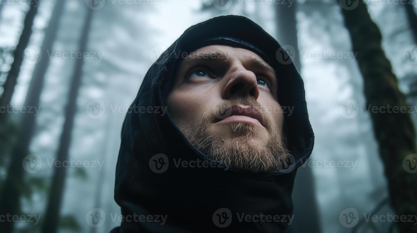 Bearded man in hooded jacket gazing upwards in misty forest, conveying adventure and contemplation photo