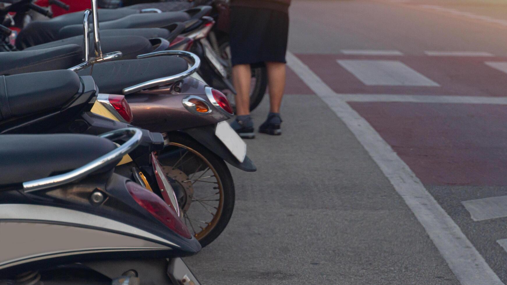 Row of parked motorcycles with a person lower legs and feet visible in the background. The scene suggests a parking area or street with parked vehicles. photo