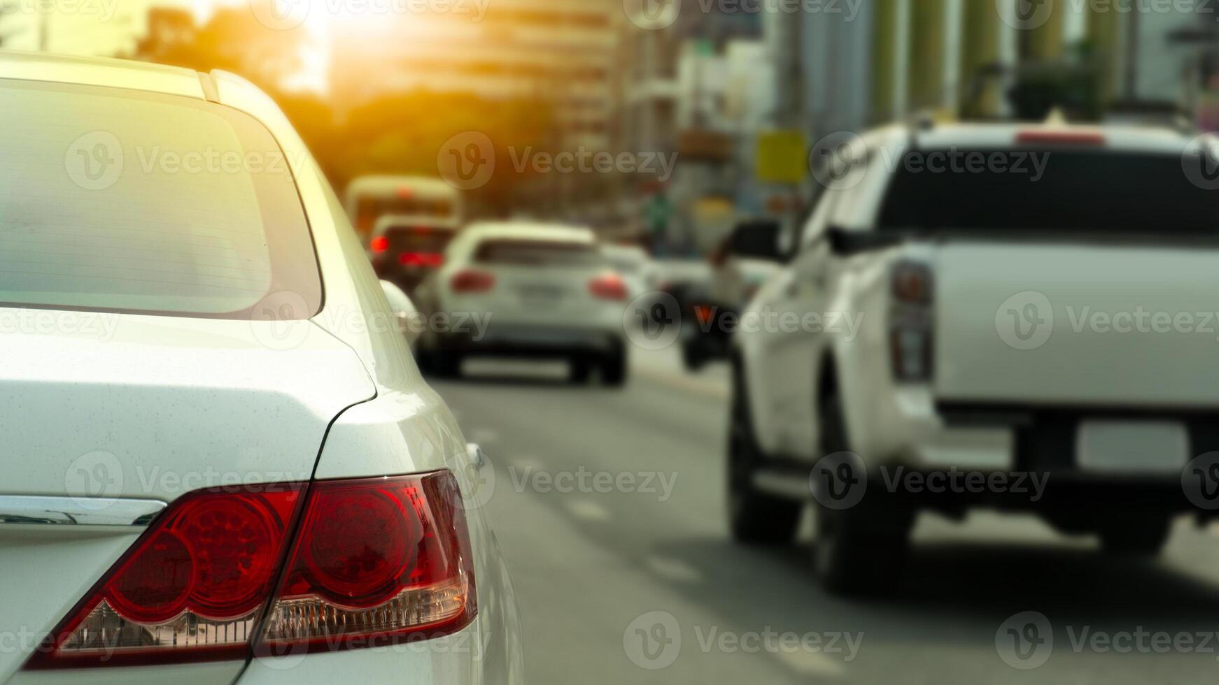 Rear side of white car driving on the asphalt road. Forward vehicular traffic. Blurred of tall buildings in the background. Light shines from behind. photo