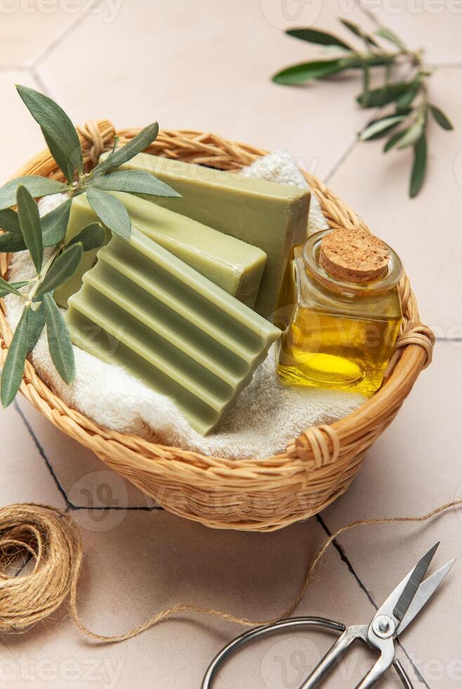 Olive soap bars, essential oil and scissors resting in wicker basket on tiled surface photo