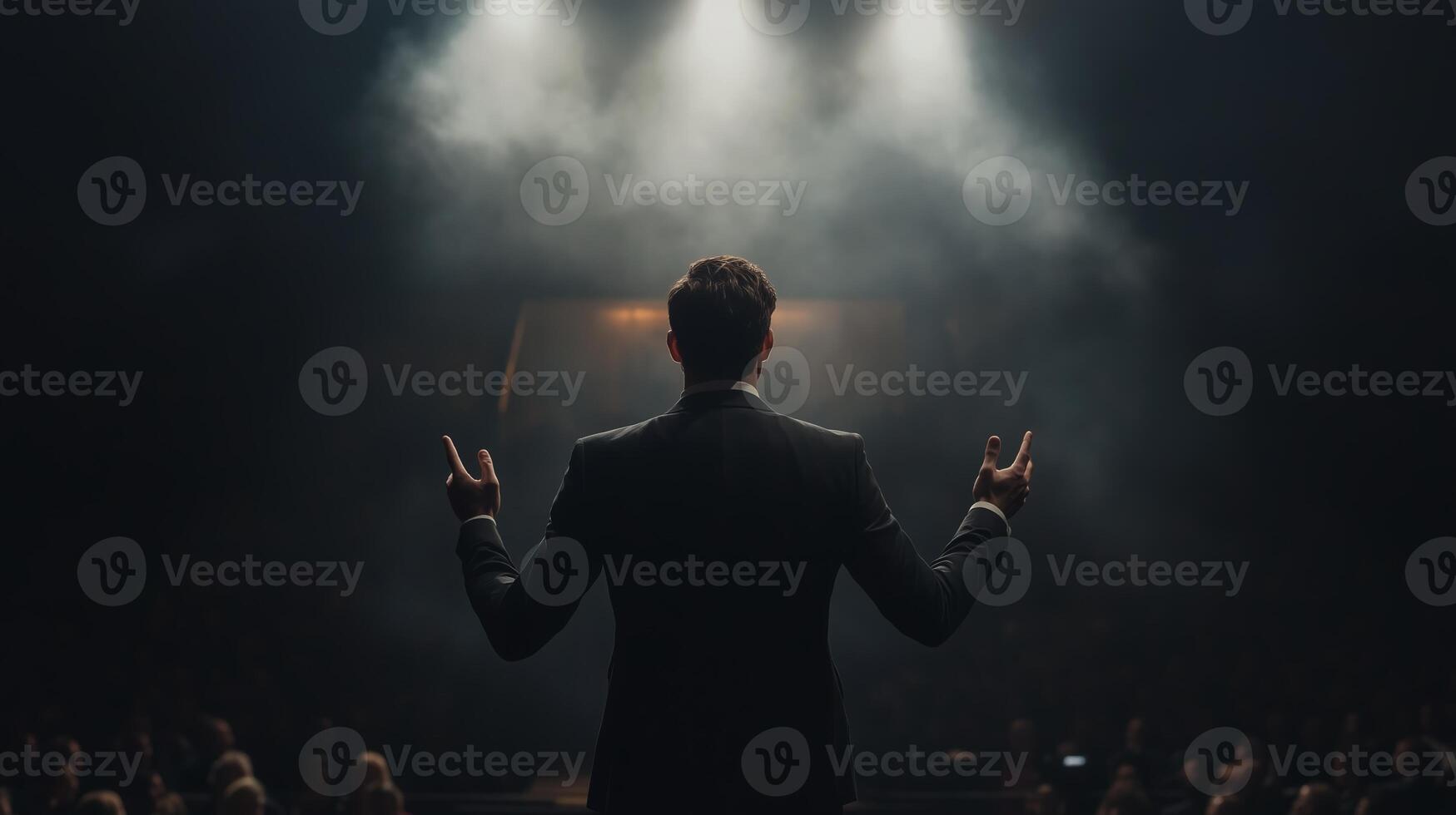 Man stands confidently on stage with hands raised, illuminated by dramatic spotlights during a performance in a theater setting photo
