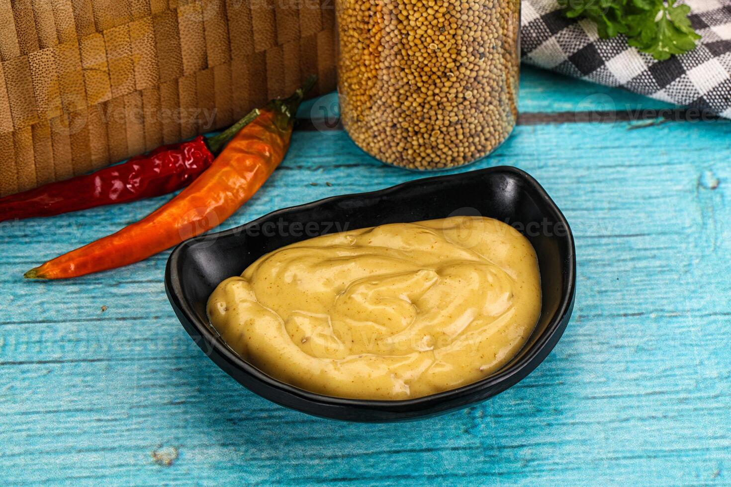 A bowl of mustard sauce with chili peppers and a basket of vegetables on a wooden table photo