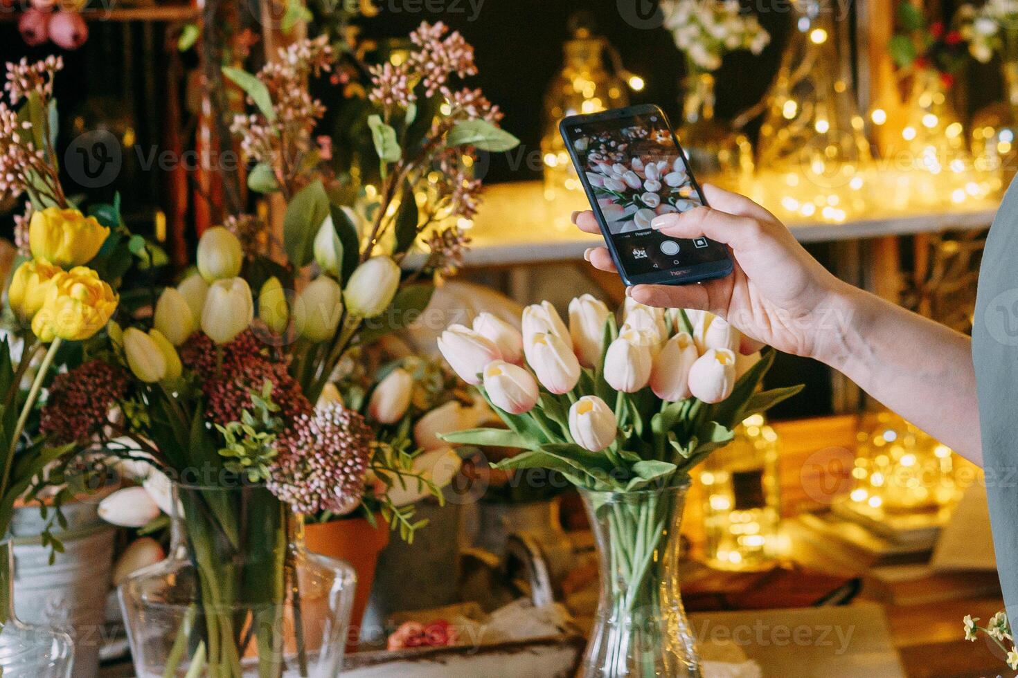 Close-up of person photographing bouquet of white tulips using smartphone in floral arrangement shop. Background features beautiful lighting and floral decor, creating warm photo
