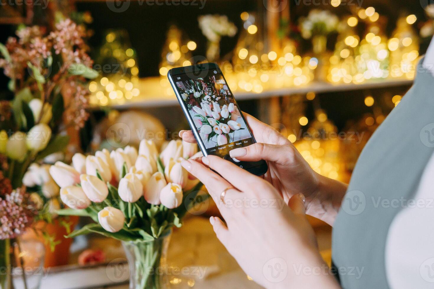 Close-up of person photographing bouquet of white tulips using smartphone in floral arrangement shop. Background features beautiful lighting and floral decor, creating warm and inviting atmosphere photo