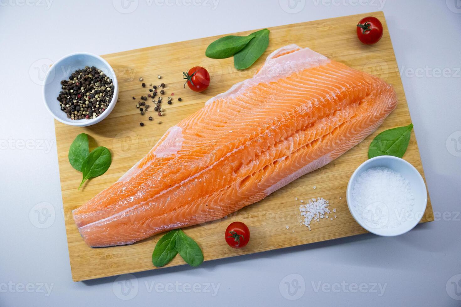 Fresh raw salmon fillet on wooden cutting board with cherry tomatoes, spinach, salt, and pepper. Healthy seafood and cooking ingredients concept. photo