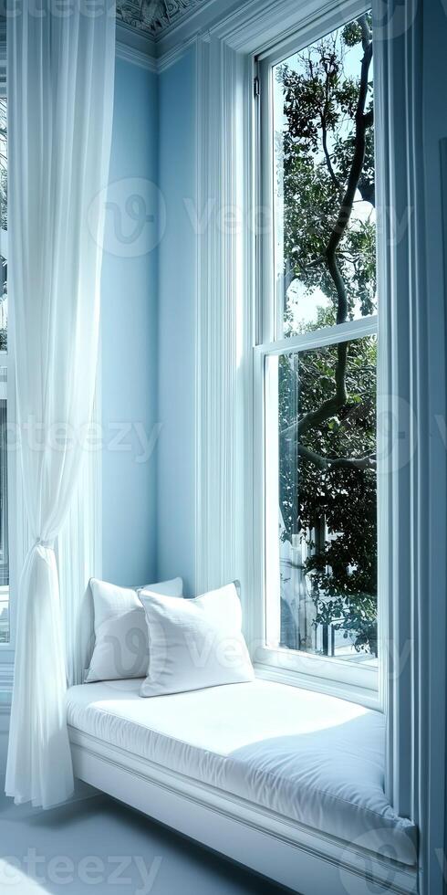 Cozy window nook featuring white cushions and soft curtains with sunlight streaming through in a serene blue room photo