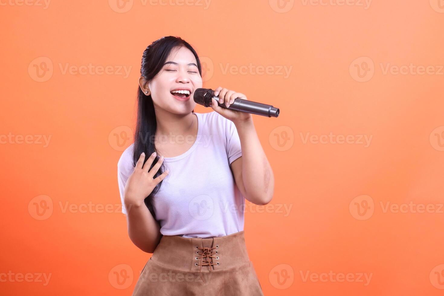 Enthusiastic young woman passionately singing into a microphone, showcasing her vocal talent and joy, isolated on orange background photo