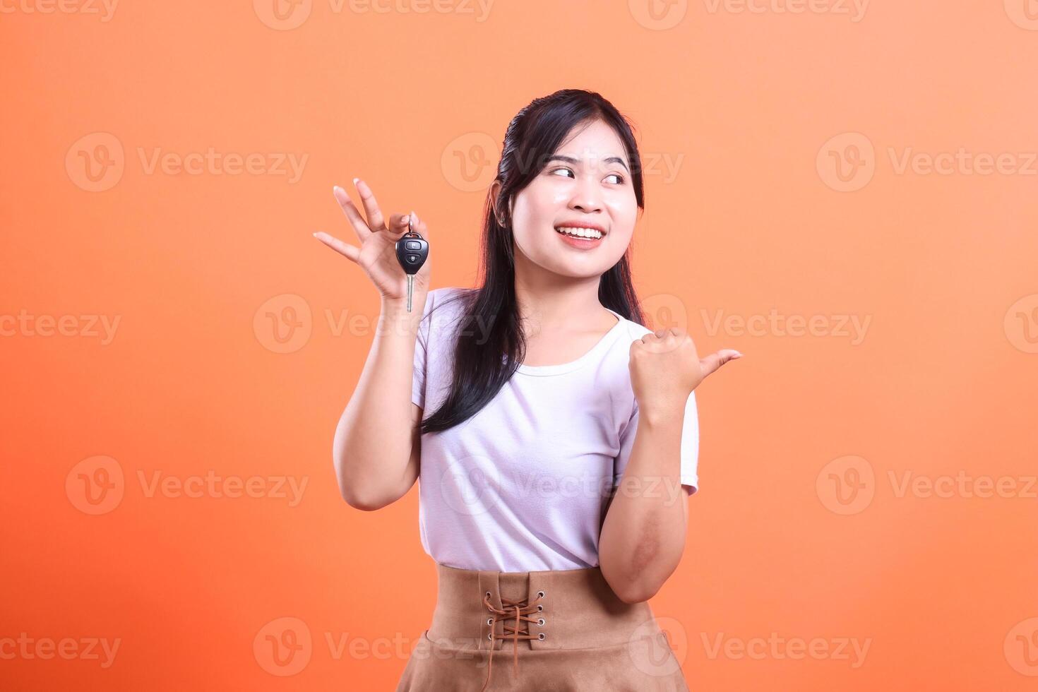 A happy Asian woman holding car keys and pointing her thumb sideways while smiling. isolated on orange background photo