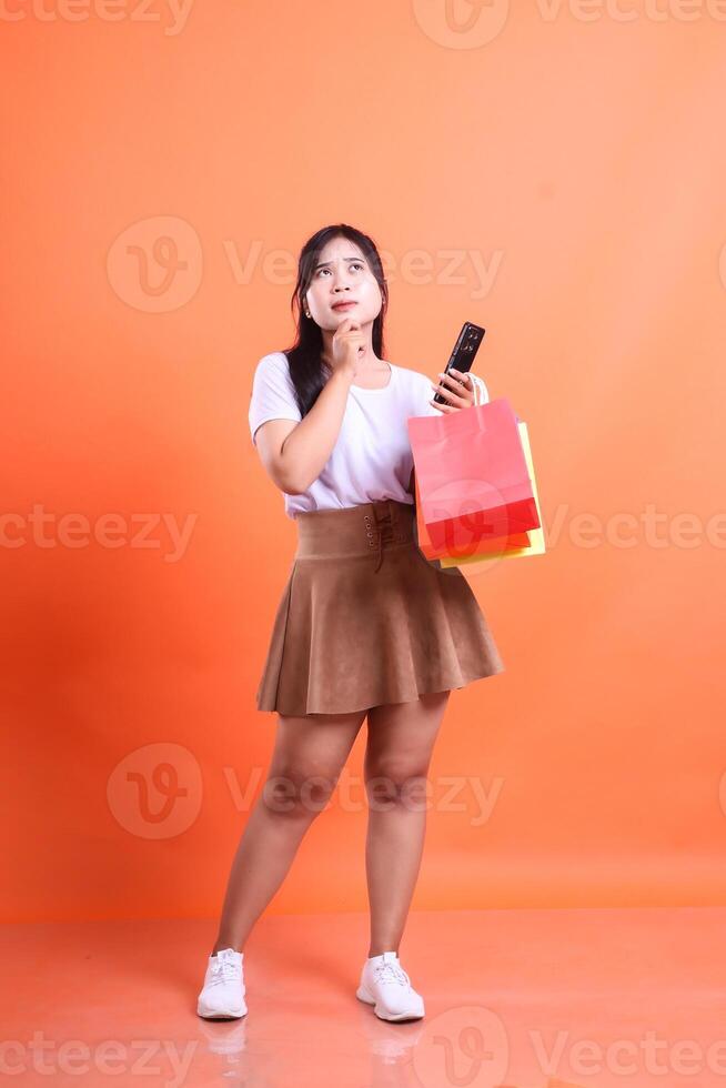 A stylish Asian woman contemplates her shopping choices, holding bags and a phone, against a bright orange background. isolated on orange background. photo