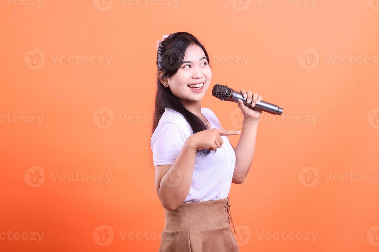 A cheerful young woman holds a microphone, smiling and pointing, ready to perform with passion. isolated on orange background photo