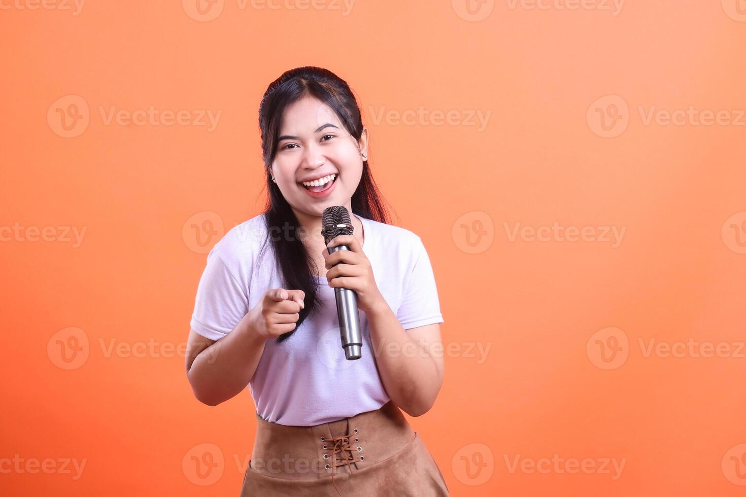 A young asian woman smiles happily while holding a microphone and pointing her finger with a positive attitude, isolated on orange background photo
