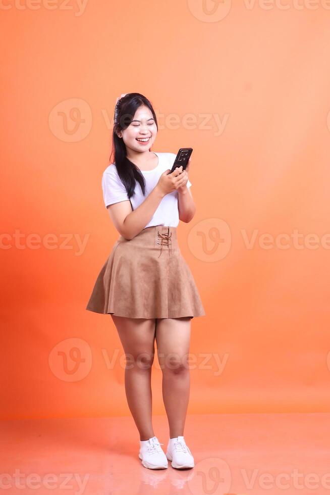 Young smiling Asian woman using smartphone in studio looking happy reading a funny message. isolated on orange background photo