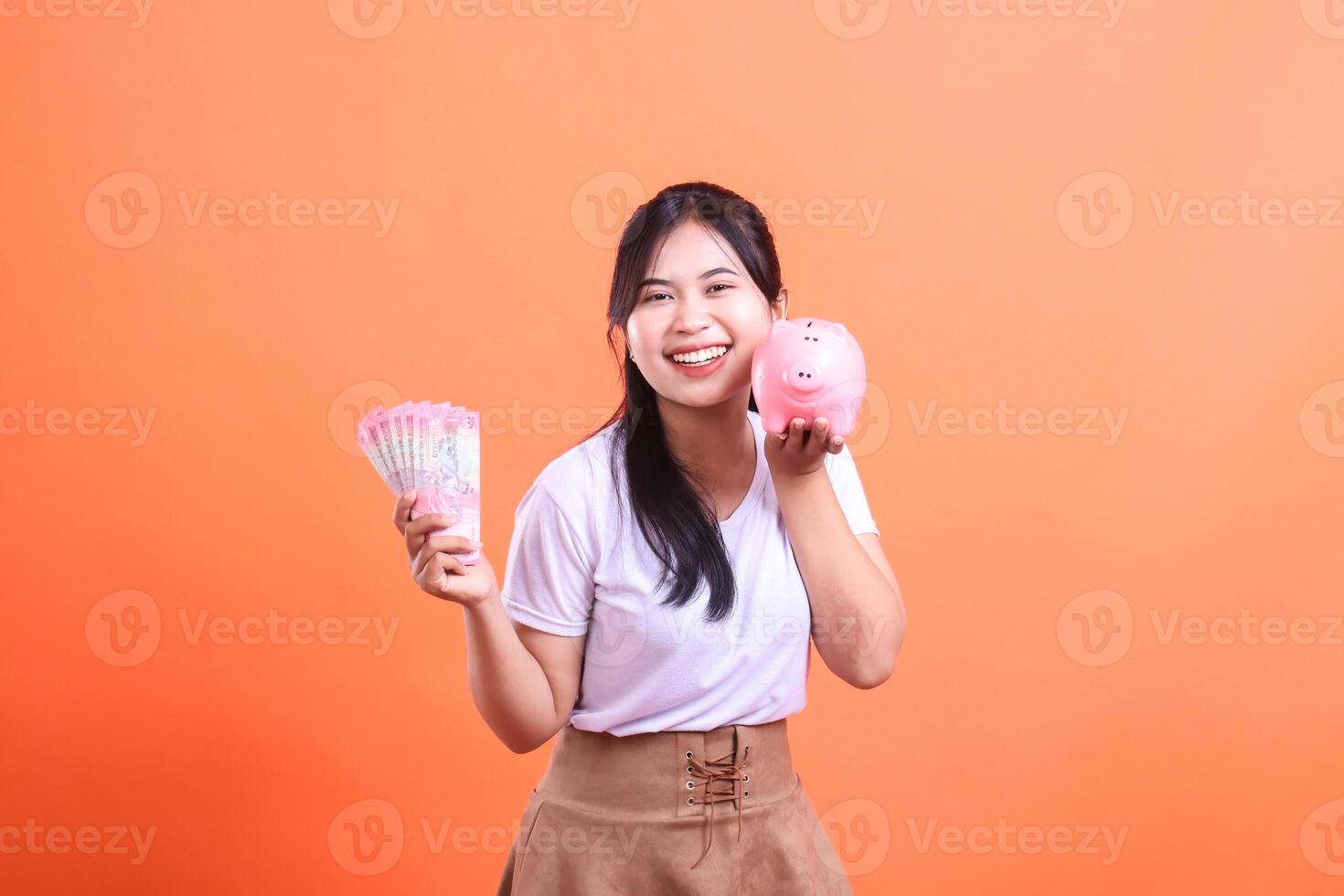 Smiling young woman holding cash and a piggy bank. Saving money for the future and investment. isolated on orange background photo