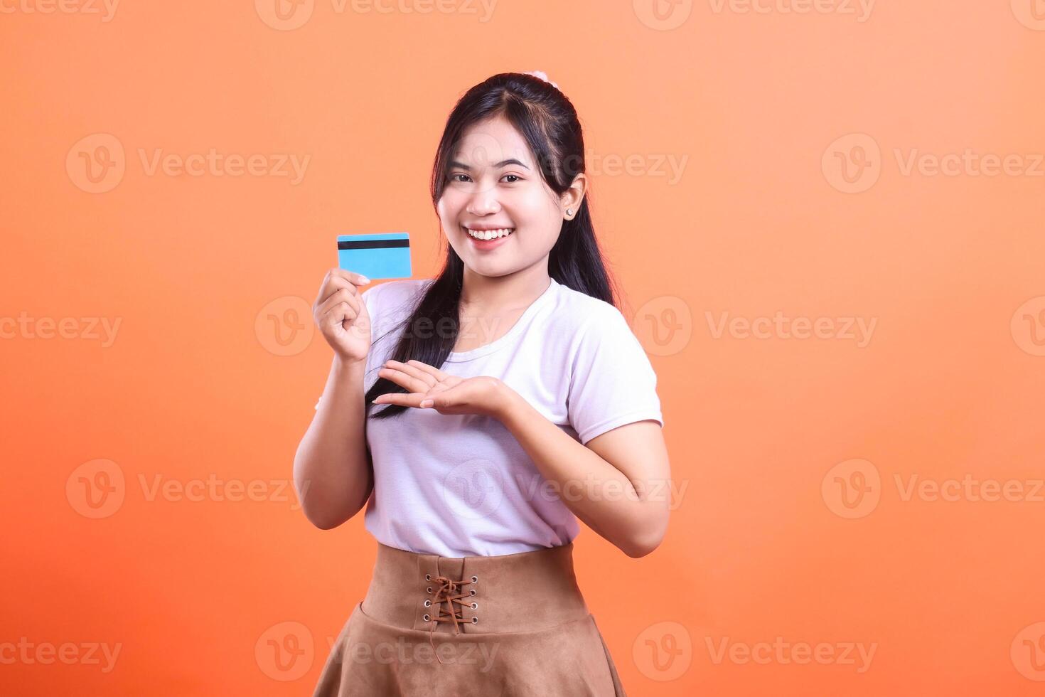 A smiling young woman presents a credit card, looking positive and confident for shopping and online purchases, isolated on orange background. photo