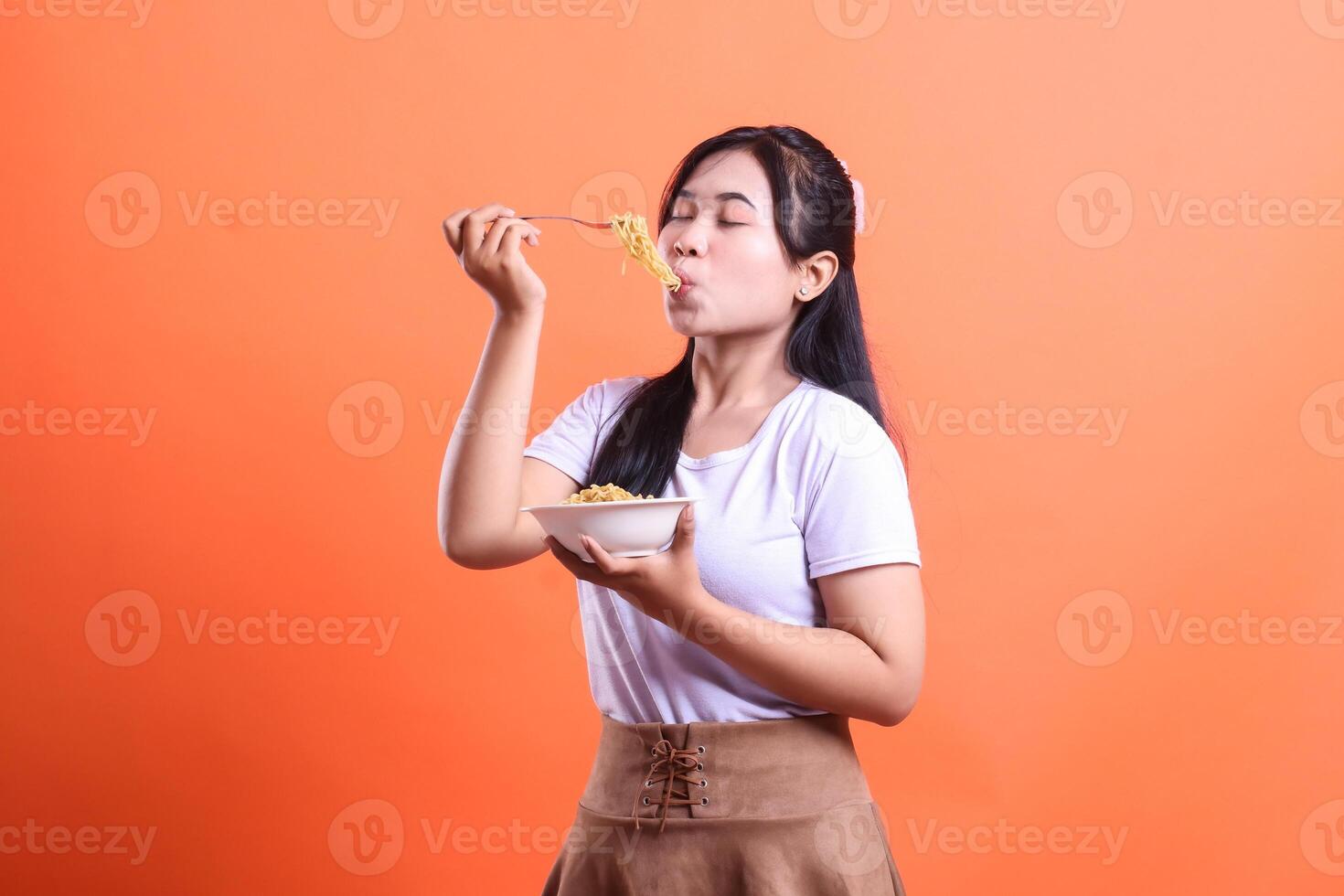 A young woman is blissfully eating her plate of noodles with her eyes closed, showing how enjoyable the meal is. isolated on orange background photo