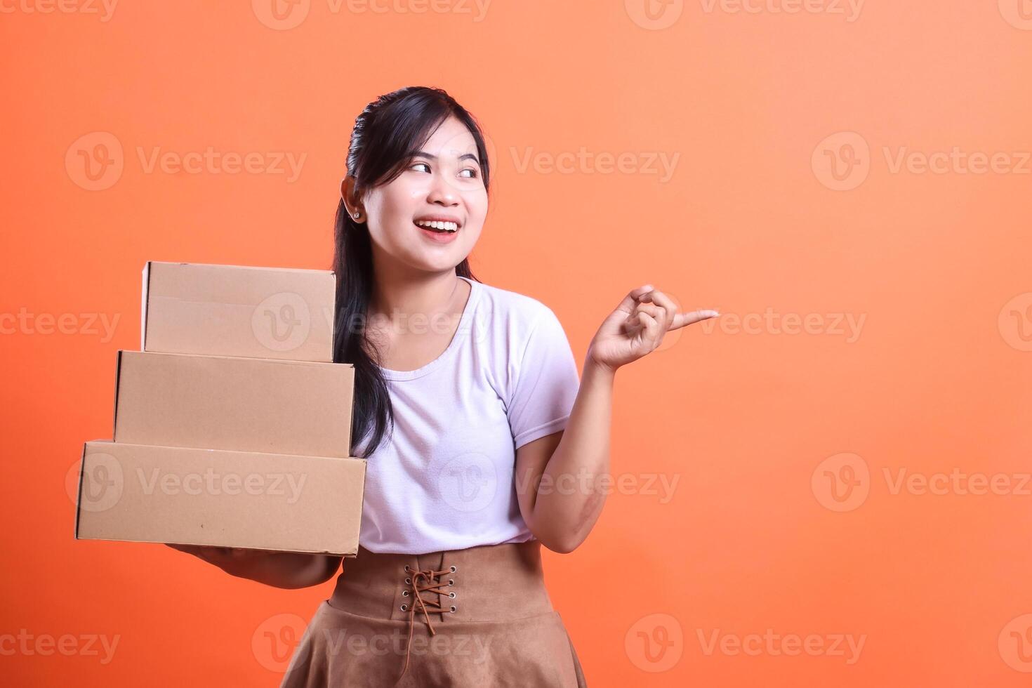 Smiling asian woman holds stacked boxes, pointing her finger towards something and looks happy. isolated on orange background photo