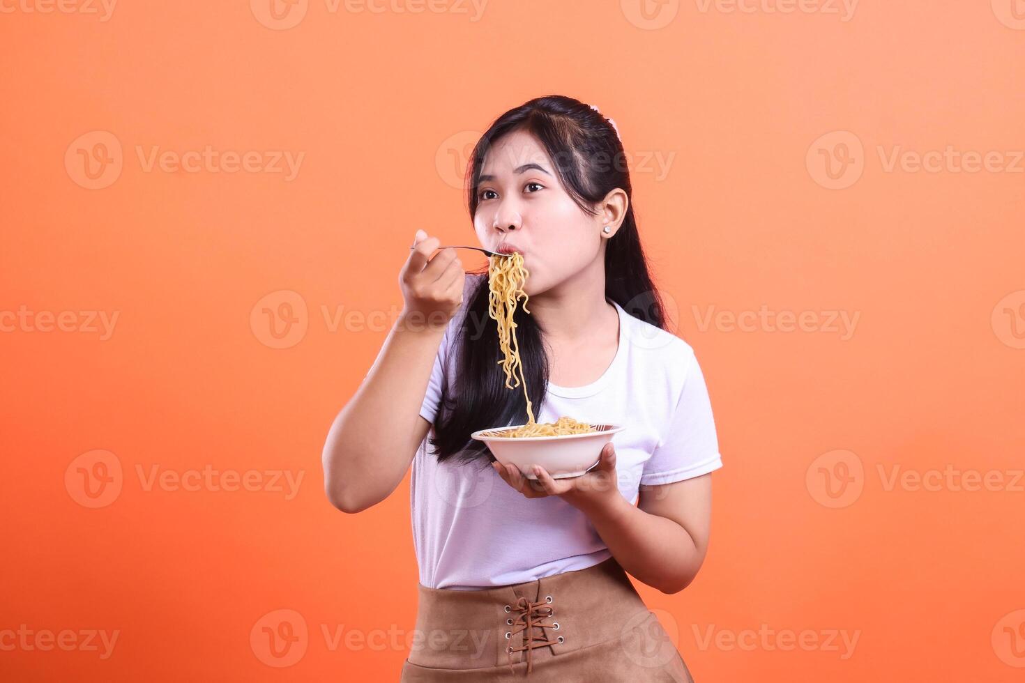 A beautiful young woman is eating noodles from a bowl with a fork, enjoying a tasty meal, isolated on orange background photo